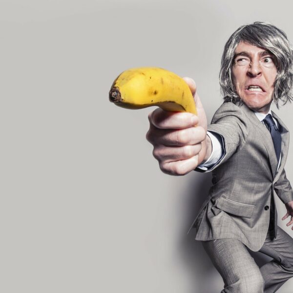 Mann mit Perücke hält eine Banane wie ein Pistole und macht eine Grimasse